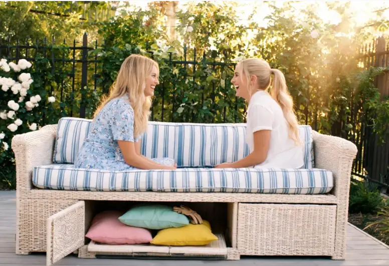 Wicker outdoor storage sofa on backyard patio with open under‑seat storage compartment holding throw pillows, two women chatting on garden furniture